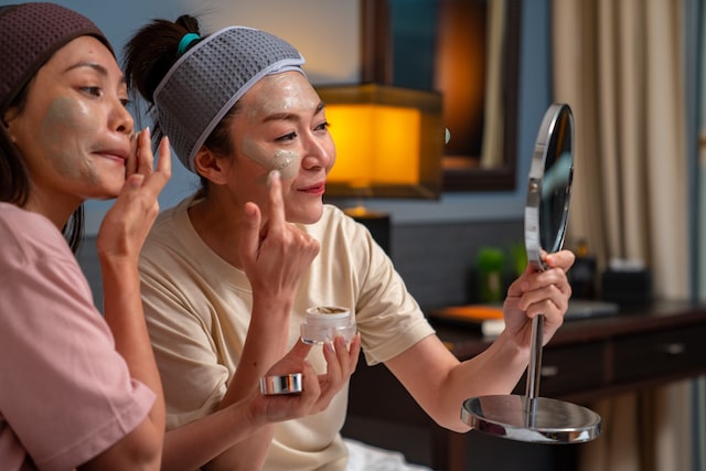 smiling asian woman friends sitting on the bed with applying skin care facial mask